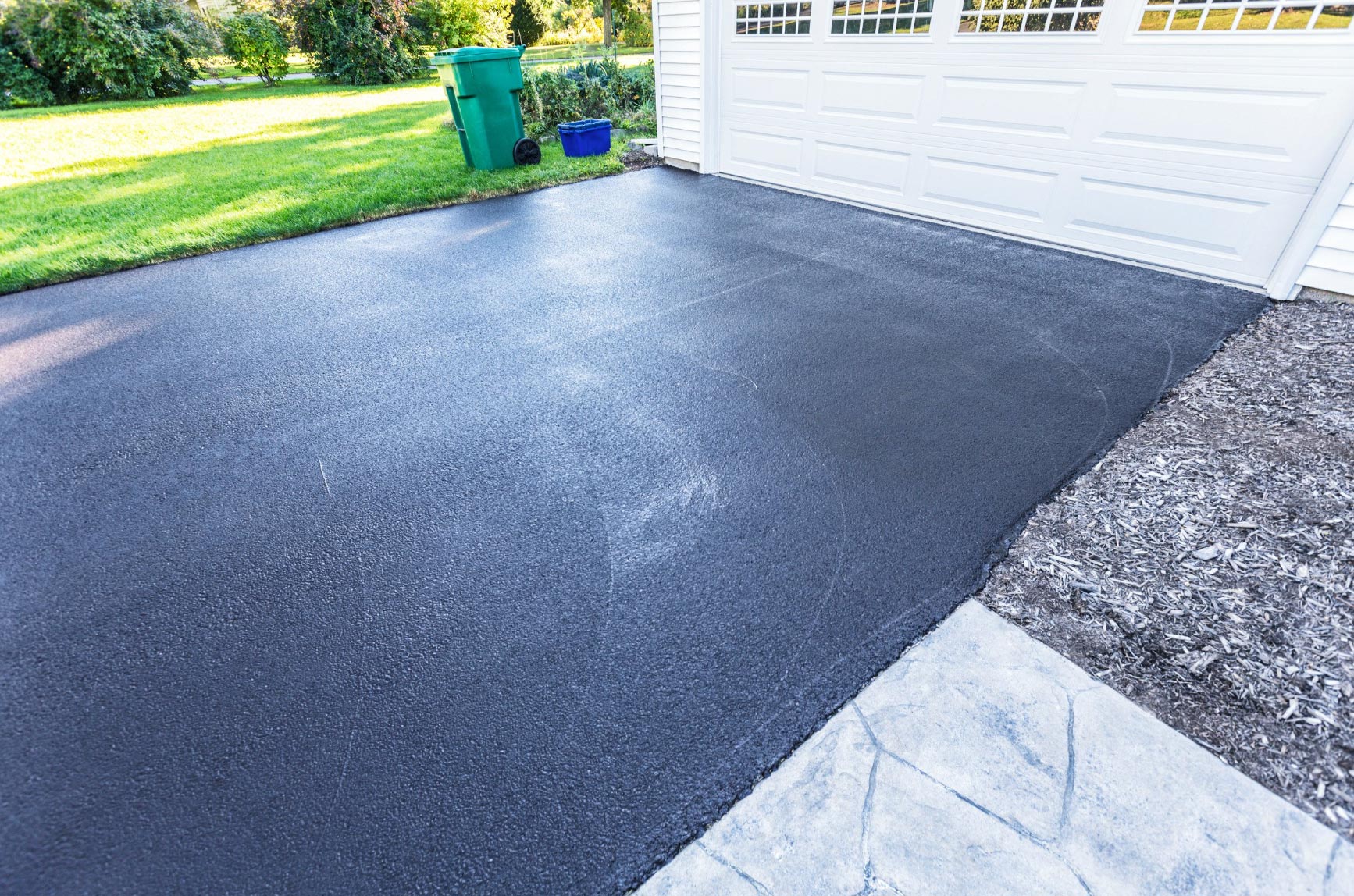 A freshly paved asphalt driveway by Will's Asphalt Paving Services sits in front of a white garage, bordered by lush lawn and landscaping.