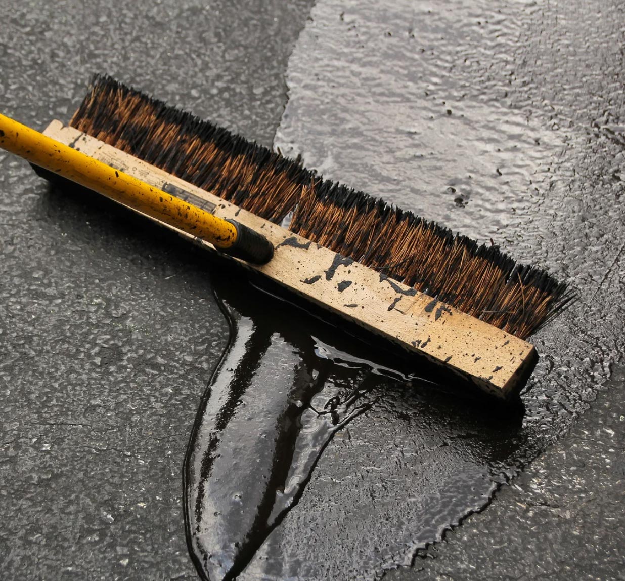 A broom with a yellow handle pushes water across a wet, textured black surface, showcasing the results of expert Newark asphalt paving services.