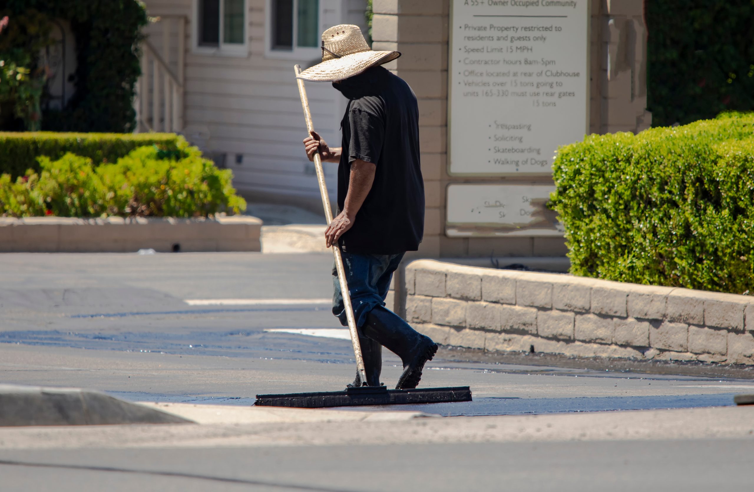A person in a wide-brimmed hat spreads asphalt on a road with a long-handled tool on a sunny day.