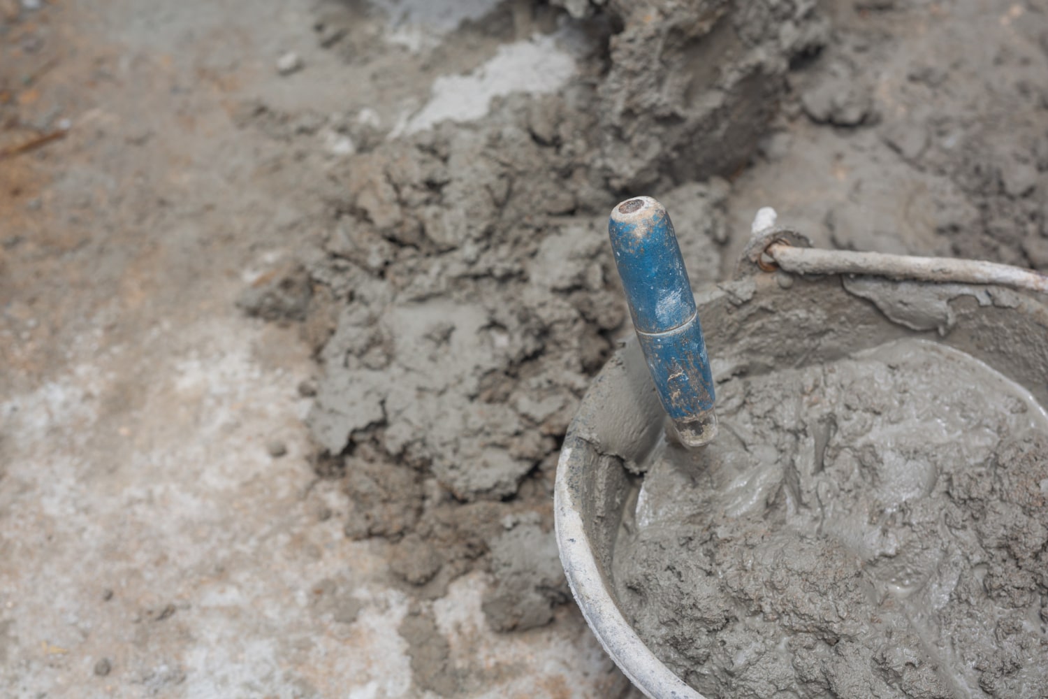 A trowel with a blue handle rests in a bucket of wet cement on a construction site.