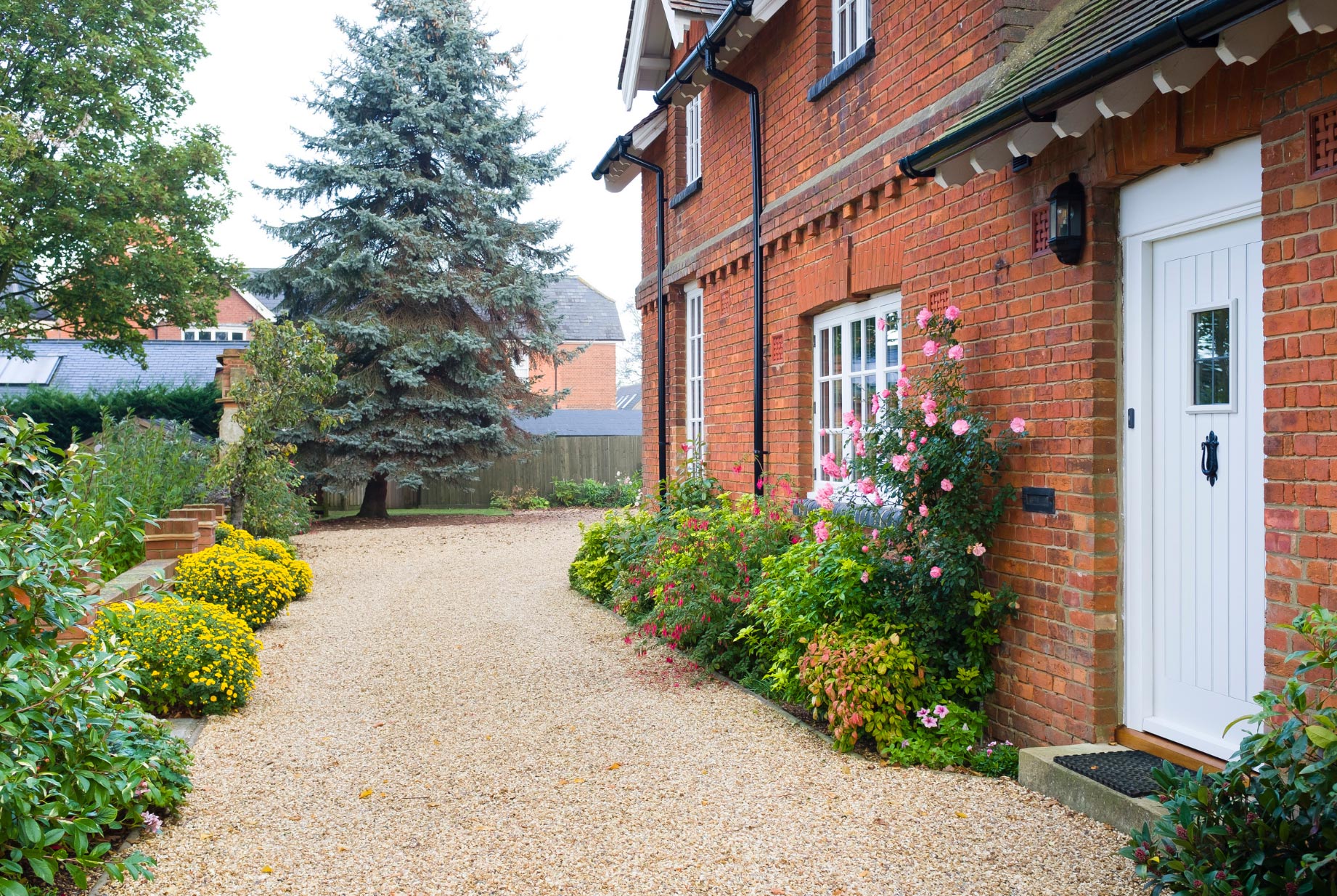 gravel-img-2 Brick house with white door, flower beds, and a gravel driveway lined with bushes and trees.